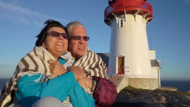 Una pareja madura admira la puesta de sol en la costa del mar con un antiguo faro . — Vídeos de Stock