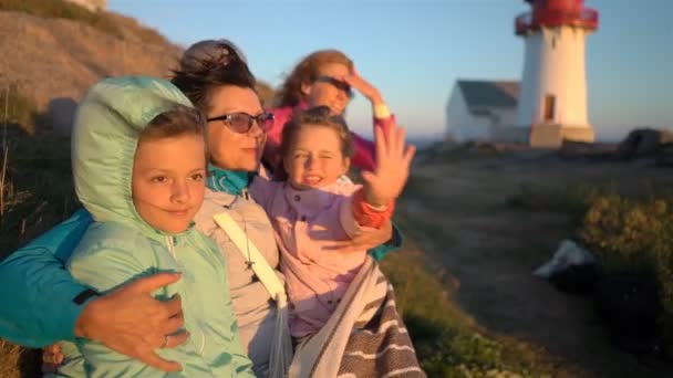 Familia feliz admirando el atardecer o el amanecer en la rocosa costa norte del mar con un antiguo faro — Vídeos de Stock