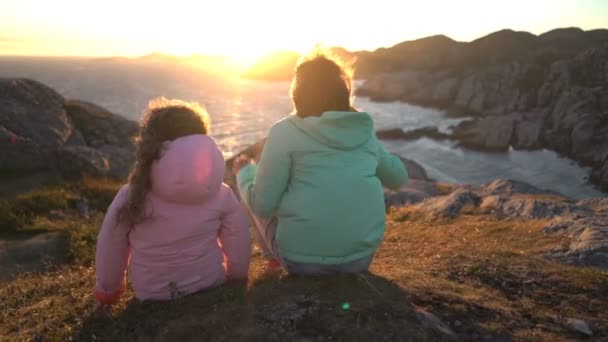 Duas meninas admiram o pôr do sol ou o amanhecer sentado nas rochas costeiras na praia do norte — Vídeo de Stock