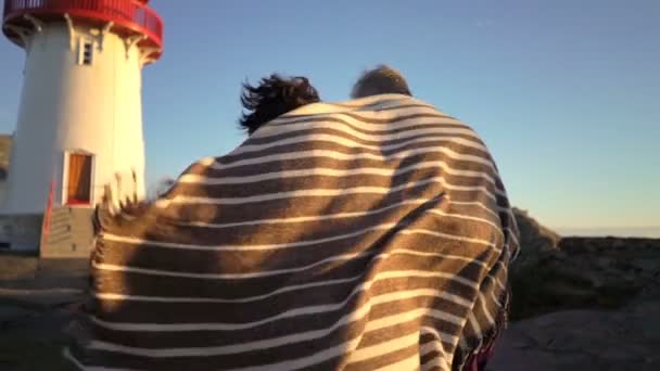 Зріла пара ходить на заході сонця на березі моря зі старим маяком — стокове відео