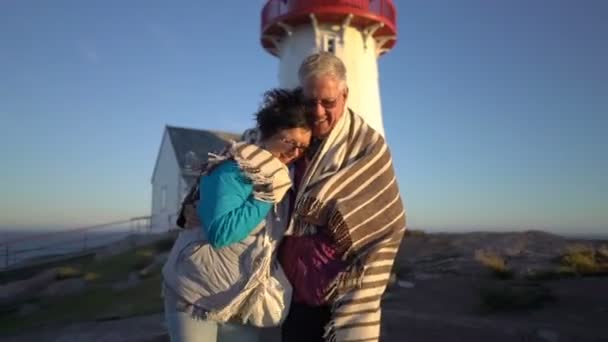 Couple d'âge mûr marchant au coucher du soleil sur le bord de la mer avec un vieux phare — Video