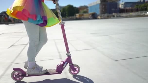 Een klein meisje in een iriserende heldere rok is het berijden van een scooter in de stad. — Stockvideo