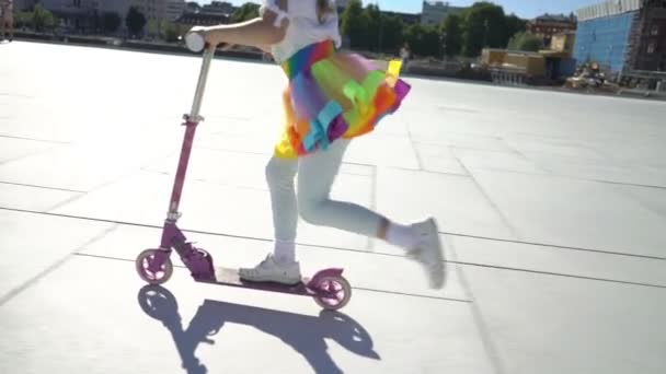 A little girl in an iridescent bright skirt is riding a scooter in the city. — Stock Video