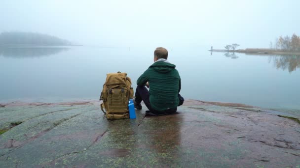 Kayalık bir Baltık Denizi kıyısında oturan sırt çantası olan orta yaşlı bir adam. — Stok video