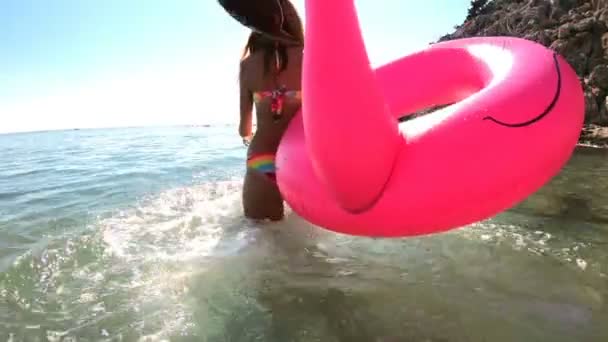 Joven mujer esbelta disfruta del mar y el sol caminando en el agua con un flamenco inflable rosa — Vídeos de Stock