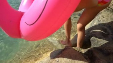 Denizin ve güneşin suya pembe bir şişme Flamingo ile Akdeniz sahilde yürüyüş zevk gökkuşağı bikini giyen genç ince kadın. Ağır çekim
