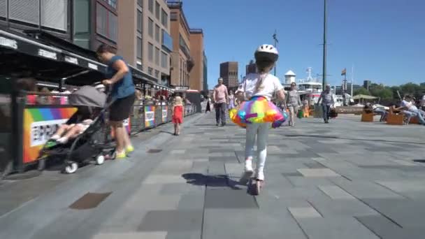 A little girl in a bright rainbow skirt and helmet rides a scooter among the city crowd in Oslo. — Stock Video