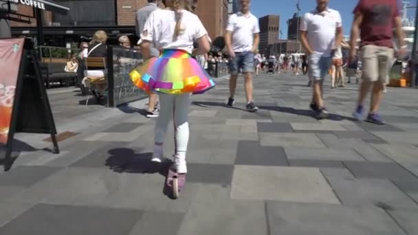 Een klein meisje in een heldere regenboog rok en helm rijdt een scooter onder de menigte van de stad in Oslo. — Stockvideo