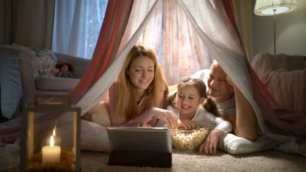 Маленькая девочка и ее родители наслаждаются просмотром мультфильмов онлайн в палатке в детской — стоковое видео