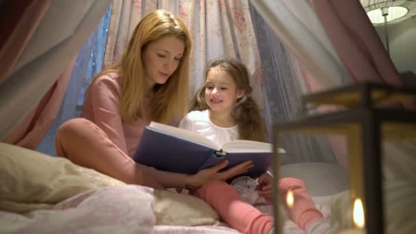 Mãe de família feliz e sua filhinha lendo um livro em uma tenda em casa — Vídeo de Stock