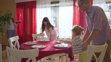 Mutlu aile evde birlikte yemek hazırlıyor, Noel ağacının yanındaki masayı parlak ışıklarla süslüyor. Sıcak tatil ortamı, aile hayatı ve kış kutlaması..