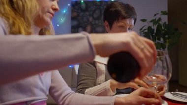 Mutlu çoklu aile Noel 'i evde kutluyor, şarap ve mum ışığında şenlikli yemek masası, bardaklara kırmızı şarap dolduruyor. Tatil atmosferi, sıcak ışıklar ve evde birliktelik kutlamaları..