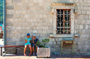 Çocuklar bir pencere bir ızgara ile eski bir taş duvarla yakınındaki bir bankta oturuyor. Ada Bogorodits kayalıkta Karadağ, Ağustos 2018