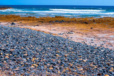 Siyah bir plajda küçük taşlar, arka planda akean dalgalar ve bir sörfçü vardır