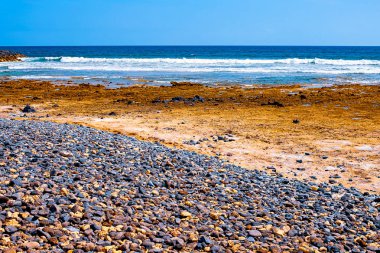 Siyah bir plajda küçük taşlar, arka planda akean dalgalar ve bir sörfçü vardır