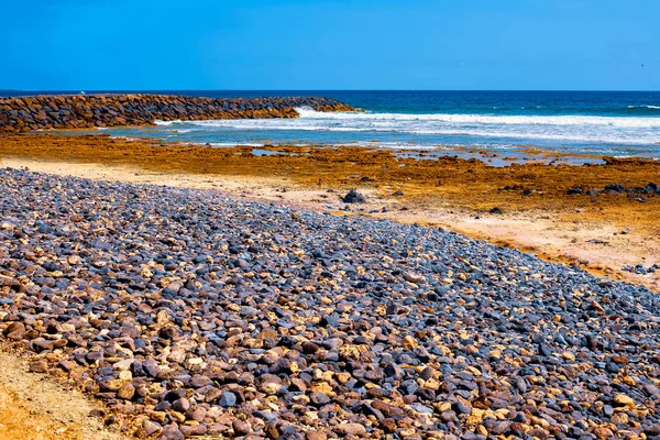 Kleine Steine auf einem schwarzen Strand, im Hintergrund akäische Wellen und ein Surfer — Stockfoto