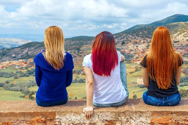 Três meninas, morena e ruiva, sentam-se de costas para a câmera contra o pano de fundo de um vale da montanha — Fotografia de Stock
