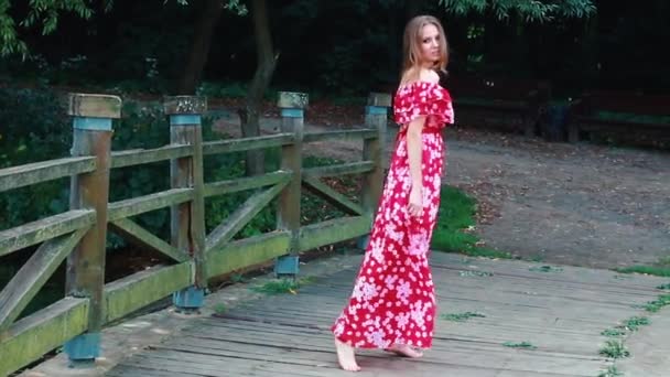 A brown-haired young girl in a red long dress walks, swirls, dances with a barefoot on a wooden bridge in a park — Wideo stockowe