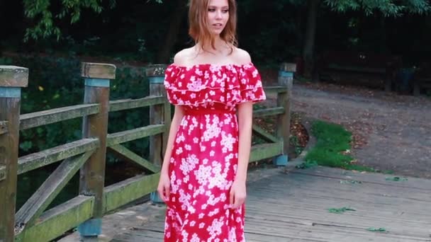 A brown-haired young girl in a red long dress walks, swirls, dances with a barefoot on a wooden bridge in a park — Vídeo de Stock