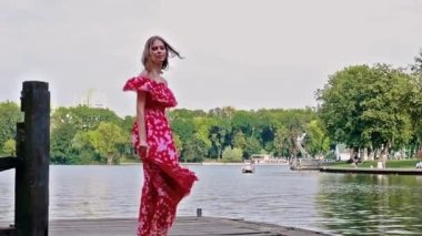 Kahverengi saçlı, uzun elbiseli genç bir kız yeşil ağaçların arka planına karşı parktaki tahta bir köprüde çıplak ayakla dans ediyor.