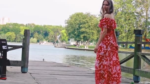 Une jeune fille brune en robe rouge longue marche, tourbillons, danse pieds nus sur un quai de bois dans un parc — Video