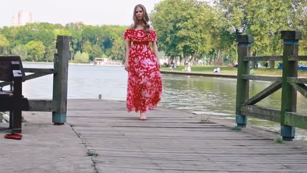 Μια νεαρή καστανή κοπέλα με ένα κόκκινο μακρύ φόρεμα περπατά, στροβιλίζεται, χορεύει ξυπόλητη σε μια ξύλινη προβλήτα σε ένα πάρκο — Αρχείο Βίντεο