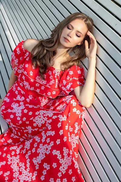 Красивая молодая девушка, модель, каштановые волосы женщина с длинными волосами в красном платье сидит на скамейке и позирует с расслабленным, вялым взглядом . — стоковое фото