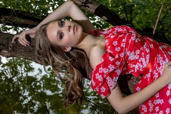 Krásná mladá dívka s perfektní pletí, s dlouhými vlasy v červených šatech leží na větvi stromu s uvolněným, malátným vzhledem. — Stock fotografie
