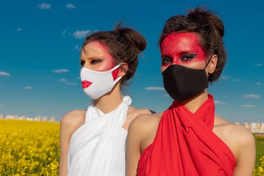 İki güzel genç esmer kız, mavi gökyüzü ve sarı çiçeklerden oluşan bir tarlanın arka planında parlak, tunik makyajlı. Tıbbi maskeli iki kız. Sağlık Hizmetleri