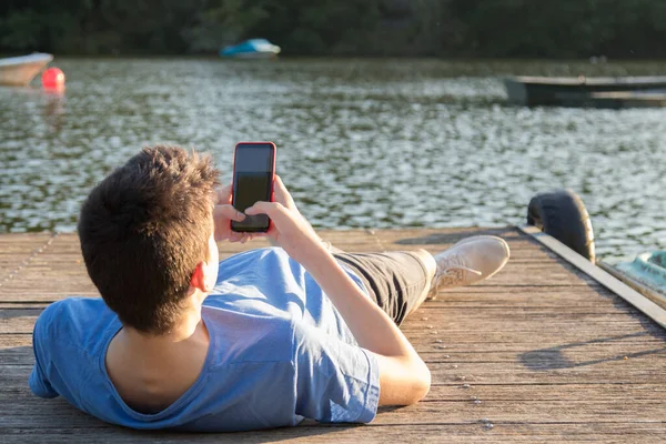 日落时带着手机坐在防波堤上的年轻人 — 图库照片
