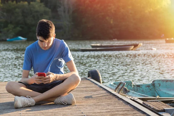 日落时带着手机坐在防波堤上的年轻人 — 图库照片