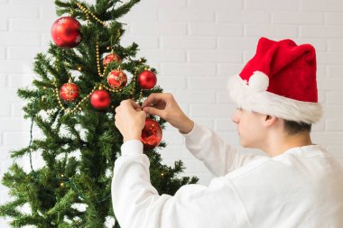 Çocuklar ön planda Noel ağacını Noel Baba şapkasıyla süslüyorlar.