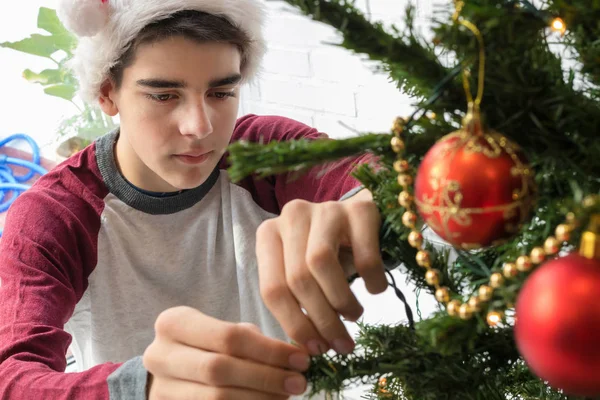 サンタ クラスの帽子をかぶった笑顔の男の子が家でクリスマスツリーを飾る — ストック写真