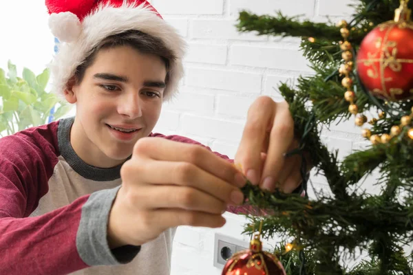 サンタ クラスの帽子をかぶった笑顔の男の子が家でクリスマスツリーを飾る — ストック写真