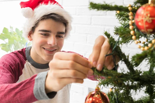 サンタ クラスの帽子をかぶった笑顔の男の子が家でクリスマスツリーを飾る — ストック写真