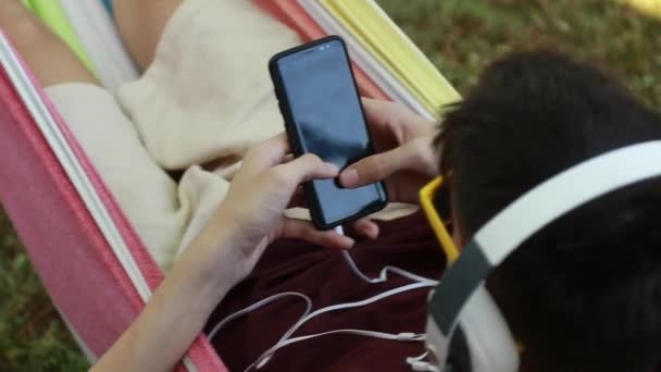 在吊床上拿着手机和耳机的年轻人 — 图库视频影像