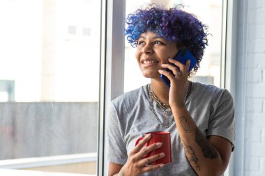 Penceresinde cep telefonu ve kahve olan Afro-Amerikalı genç bir kadın.