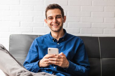 Apartmandaki koltukta oturan genç adam cep telefonuyla.