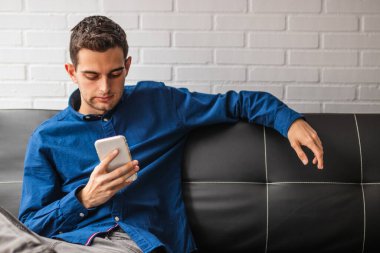 Apartmandaki koltukta oturan genç adam cep telefonuyla.