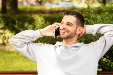 Sakallı ve cep telefonlu çekici bir genç parkta oturuyor.