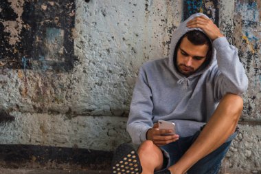 Genç adam cep telefonuyla sokak zemininde oturuyor.