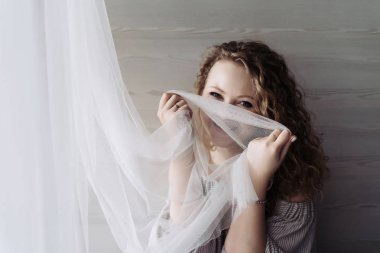 kıvırcık mutlu kadın pencerenin kenarında oturur, perdeli ve güler yüzünü kapatır