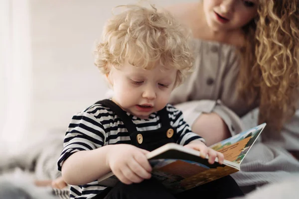 Curly little baby boy sitting on the bed and reading a book, along with mom — Stock Photo, Image