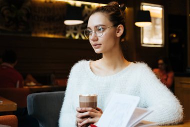 güzel genç kız öğrenci bir beyaz ceket ve gözlük okuldan sonra bir kafede oturuyor ve lezzetli kahve içiyor