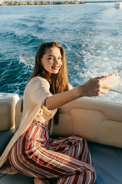 Szczęśliwa młoda dziewczyna w lato stylowe ubrania siedzi na jej biały jacht, pływa na błękitne morze i sprawia, że selfie w swoim smartfonie — Zdjęcie stockowe
