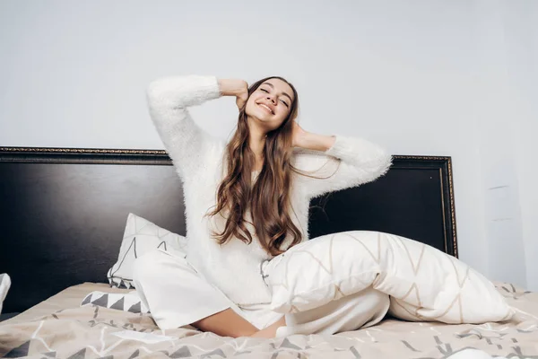 La encantadora chica de pelo largo en pijama se despertó temprano en la mañana en un buen estado de ánimo, se extiende y sonríe al nuevo día — Foto de Stock