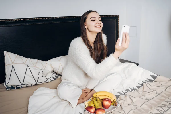 Niña feliz en pijama sentada en la cama temprano en la mañana, comiendo frutas y mirando en su teléfono inteligente — Foto de Stock