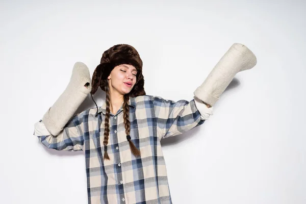 Divertida chica rusa joven con coletas y en un sombrero de piel caliente, el aura espera el invierno, mantiene las botas de fieltro gris — Foto de Stock