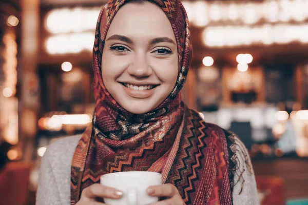 Счастливая молодая мусульманка с платком на голове пьет ароматный кофе в уютном восточном кафе и улыбается — стоковое фото