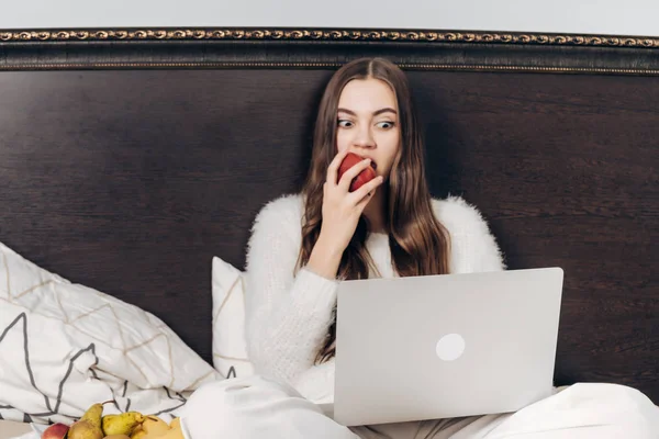 Захоплена молода дівчина сидить у ліжку ввечері і дивиться цікаве телешоу на її ноутбук їсть яблуко — стокове фото